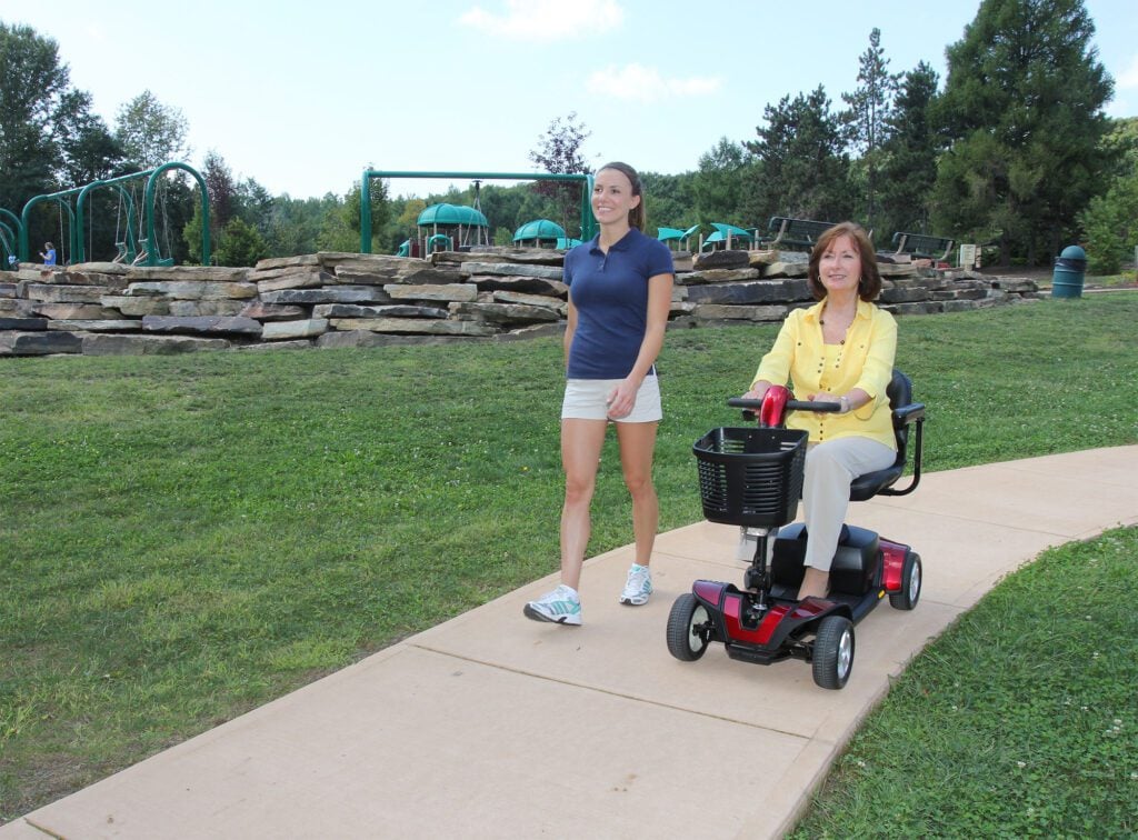 Woman riding a red mobility scooter on a park path beside a walking companion