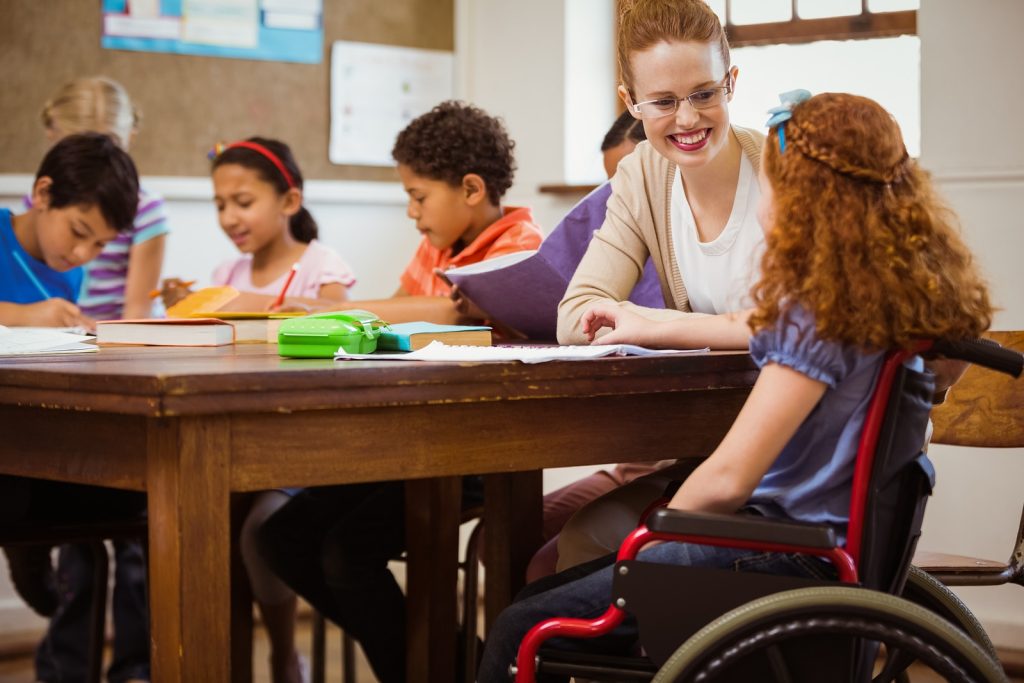 Teacher smiling with a young girl in a wheelchair at a classroom table with other students
